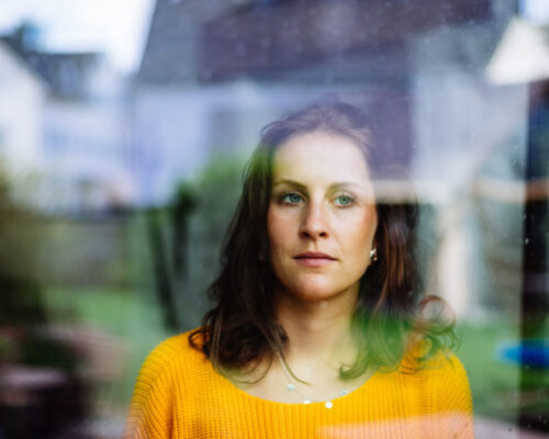 Young woman looks thoughtfully and sadly through the window into the garden with children's toys.