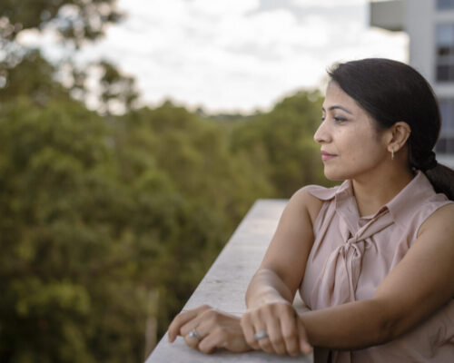 A woman of Indian descent stands on the patio of her apartment and looks thoughtfully into the distance.