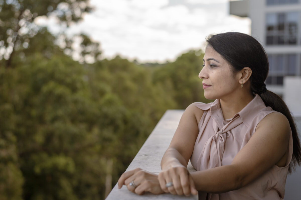 A woman of Indian descent stands on the patio of her apartment and looks thoughtfully into the distance.