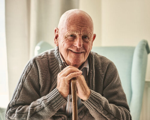 Portrait of happy senior man sitting at home with walking stock and smiling